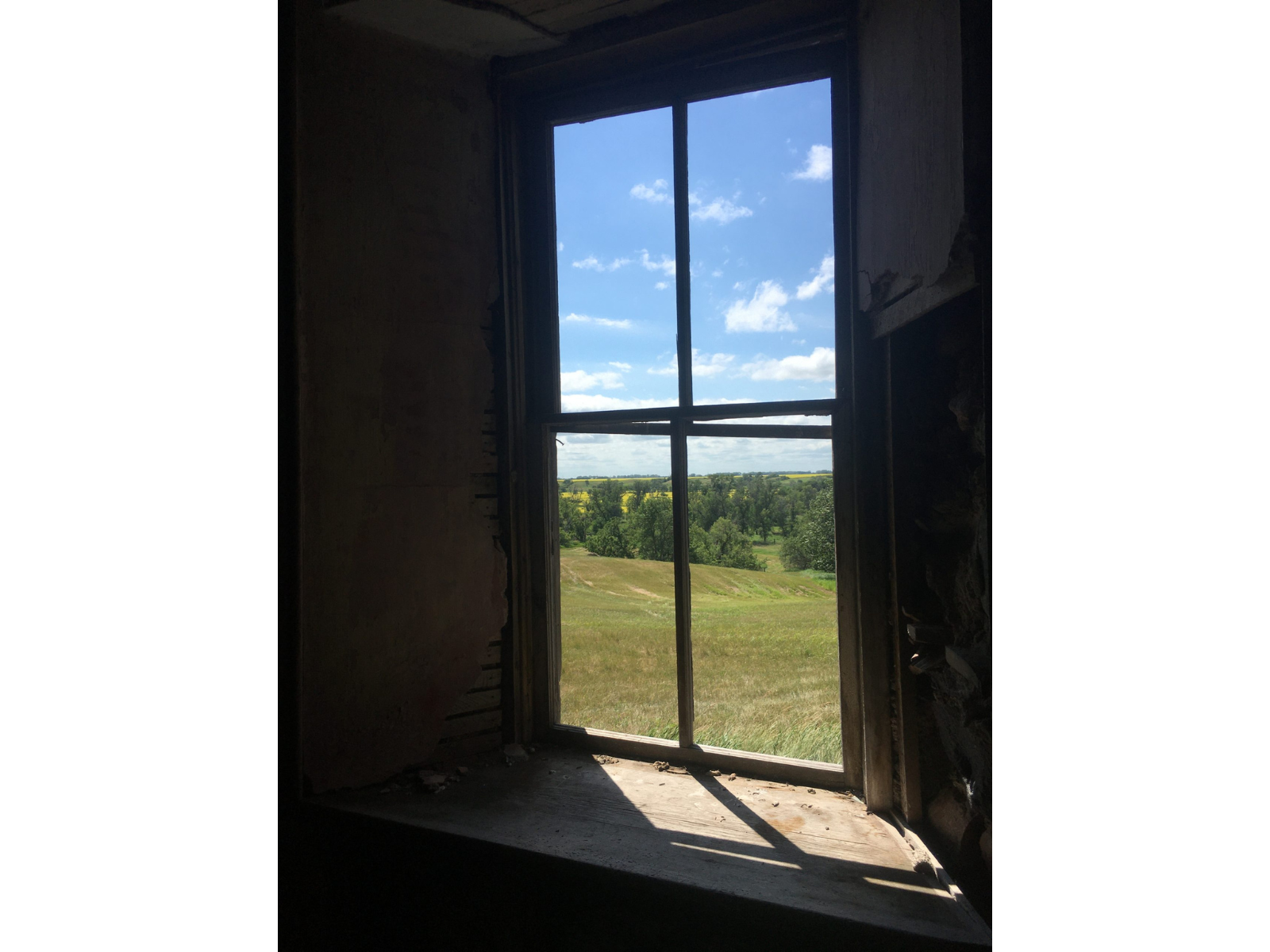 View looking out an old window, with the wooden posts framing where the panes once sat. Out the window is a view over rolling grass and green trees below a blue sky speckled with white clouds.