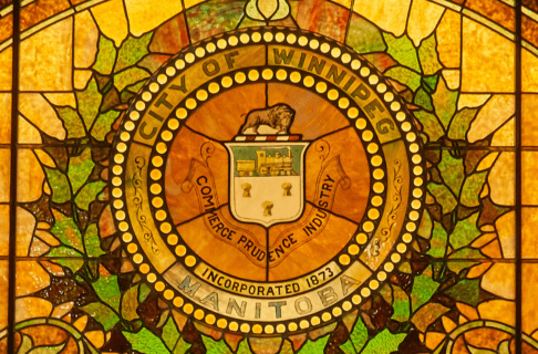 Half circle stained glass window featuring a leaves and flowers pattern in oranges and greens. In the centre, surrounded by a laurel, is the city of Winnipeg crest with text reading, "City of Winnipeg Manitoba / Incorporated 1873 / Commerce, Prudence, Industry".