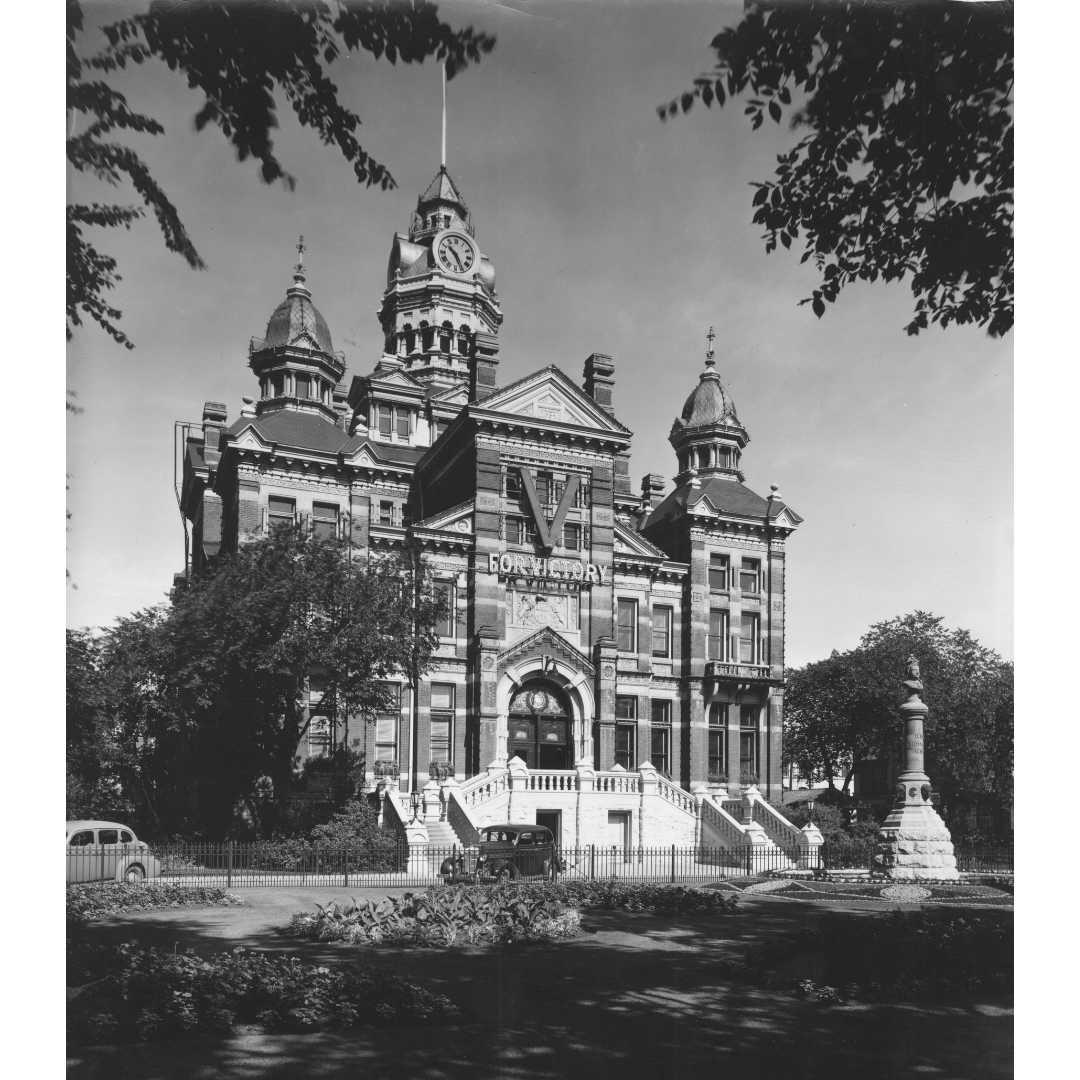 A black and white photograph of a square Victorian building with towers and turrets. Hanging on the front of the building is a large "V" with a banner reading "For Victory" beneath. Over the door below this is a semi-circle stained glass window.