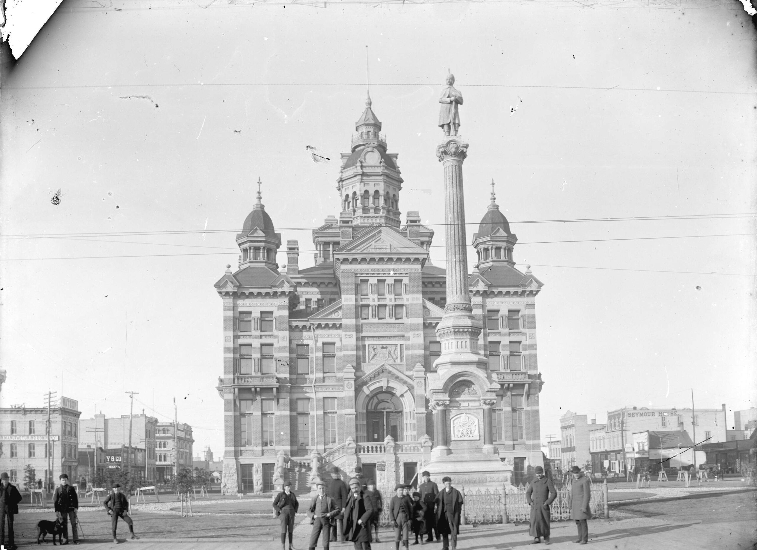 A black and white photograph of a square Victorian building with towers and turrets. A few people are grouped in the foreground, near a statue on a tall pillar.