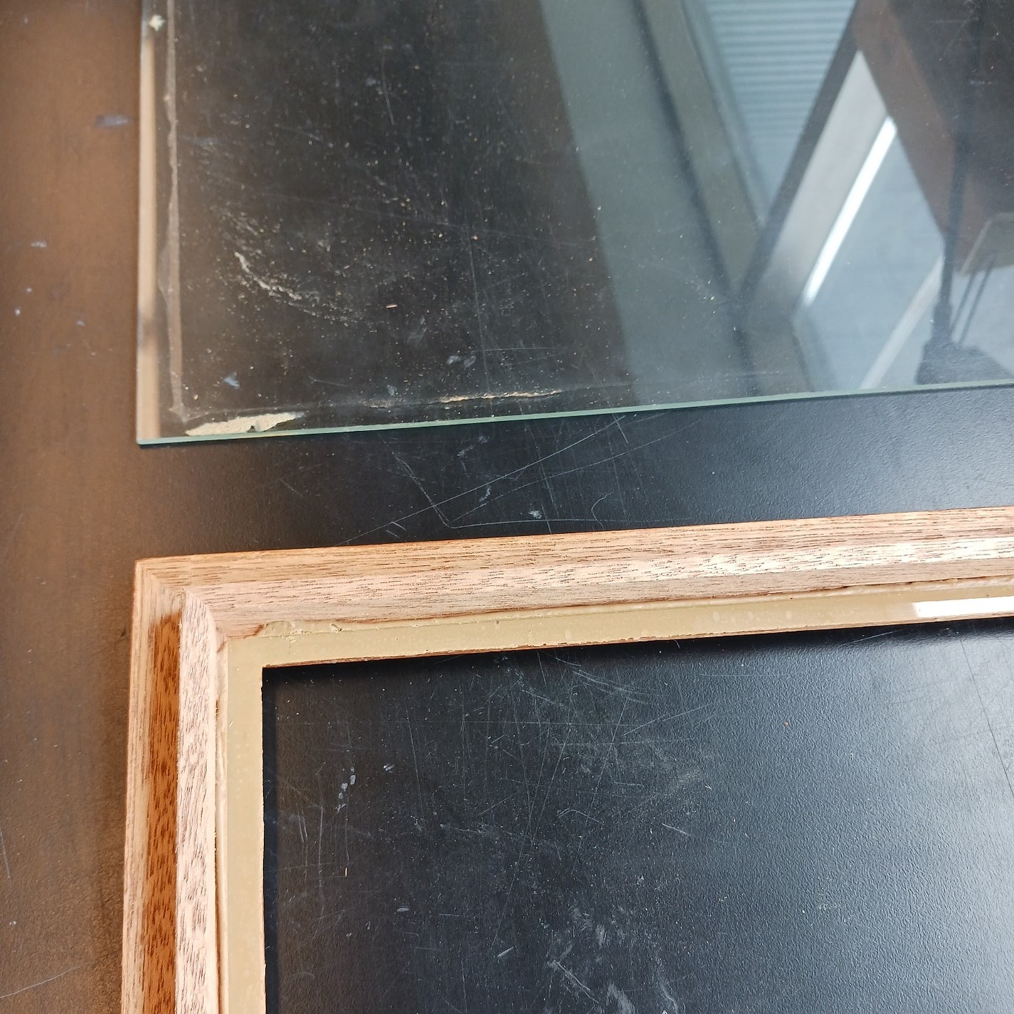 Square sheet of clear glass and broken wooden frame lying flat on a dark surface.