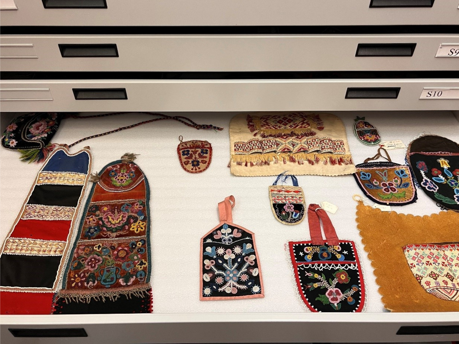 An open drawer containing twelve intricately beaded and quilled wall pockets and bags, laid out carefully for storage.