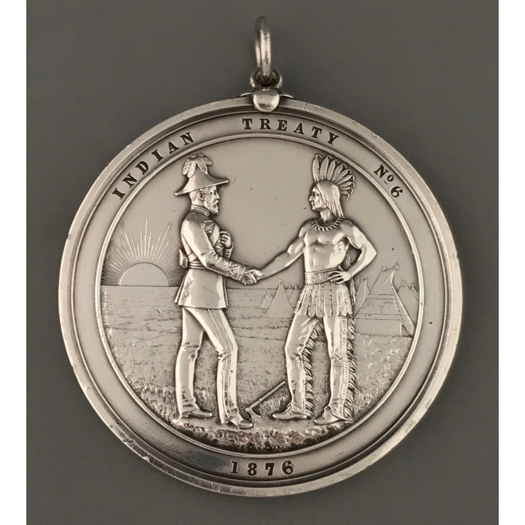 Photograph of the Treaty Number 6 handshake medal. A circular medal portraying a representative of England shaking hands with an Indigenous leader. They stand on grassy ground in front of tipis and the rising sun. Text around the edge of the medal reads, “Indian Treaty No. 6 / 1876”.