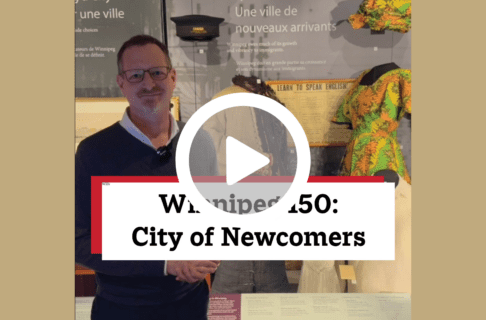 A screenshot of a video, an individual standing in front of a display case containing a number of artifacts including a dress and hat in a brightly coloured fabric. There's a play button over the screenshot and overlaid text reads, "Winnipeg150: City of Newcomers".