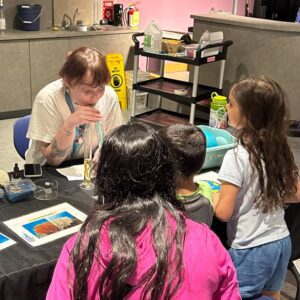 A Museum staff member performs a science experiment at a table as three children watch.