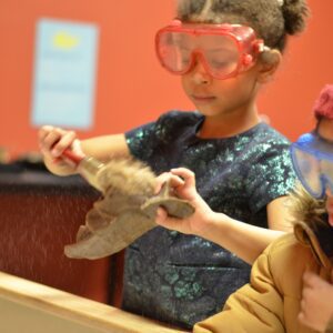 A child wearing goggles using a paintbrush to brush sand of off a fossil.