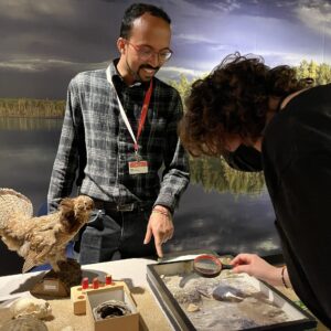 A smiling Manitoba Museum staff member points something out at an interactive display table to another individual who is leaning over to look at something through a magnifying glass.