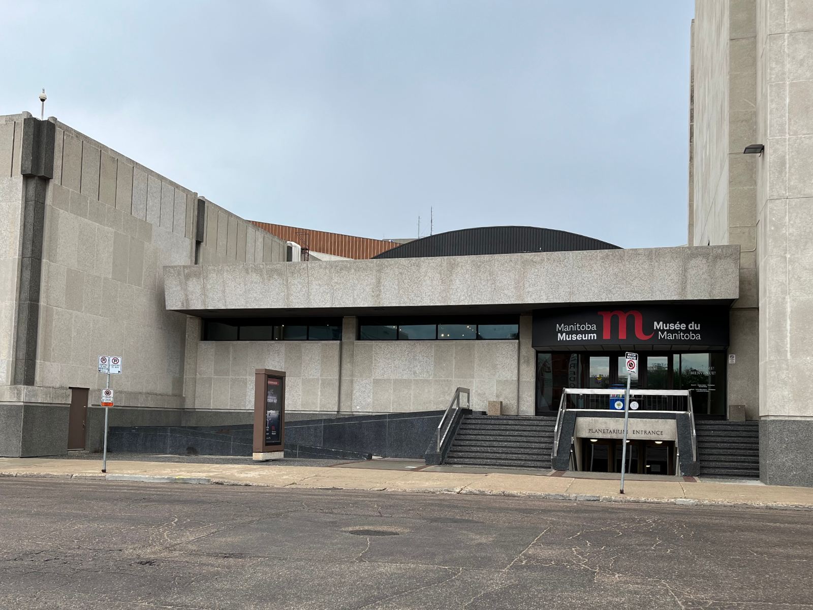 The exterior of the Rupert entrance to the Manitoba Museum in June 2024. Tyndall stone walls form the exterior wall next to a low cement ramp and stairways to the doors. 