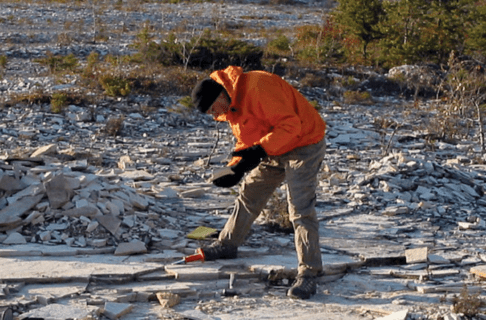 A person wearing a bright orange jacket and black toque bends over to examine a piece of stone they've picked up off the stony ground where they stand.