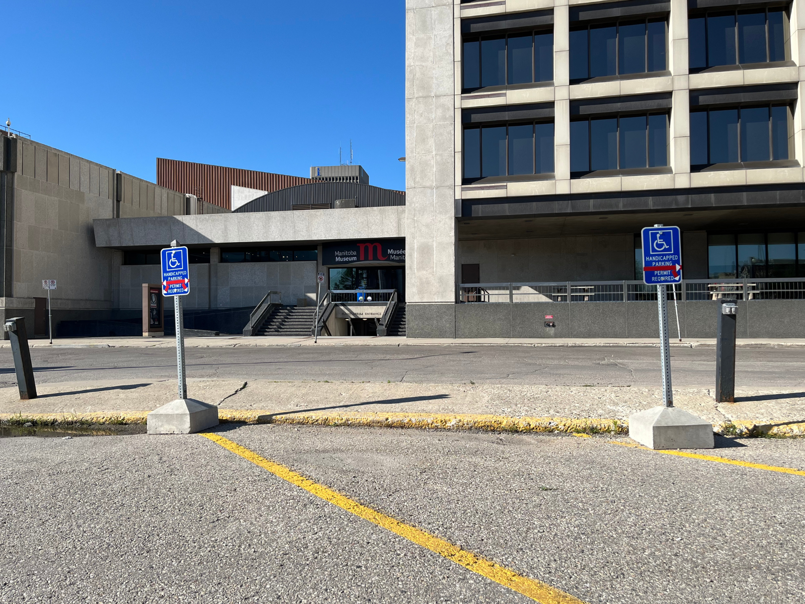 Photograph looking at the Manitoba Museum Rupert entrance from the parking lot. Two signs identify three handicapped parking spaces.