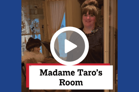 A screenshot of a video, an individual standing in a Museum exhibit - a space set up like a bedroom with a mannequin seated at a small table.. There's a play button over the screenshot and overlaid text reads, "Madame Taro's Room".