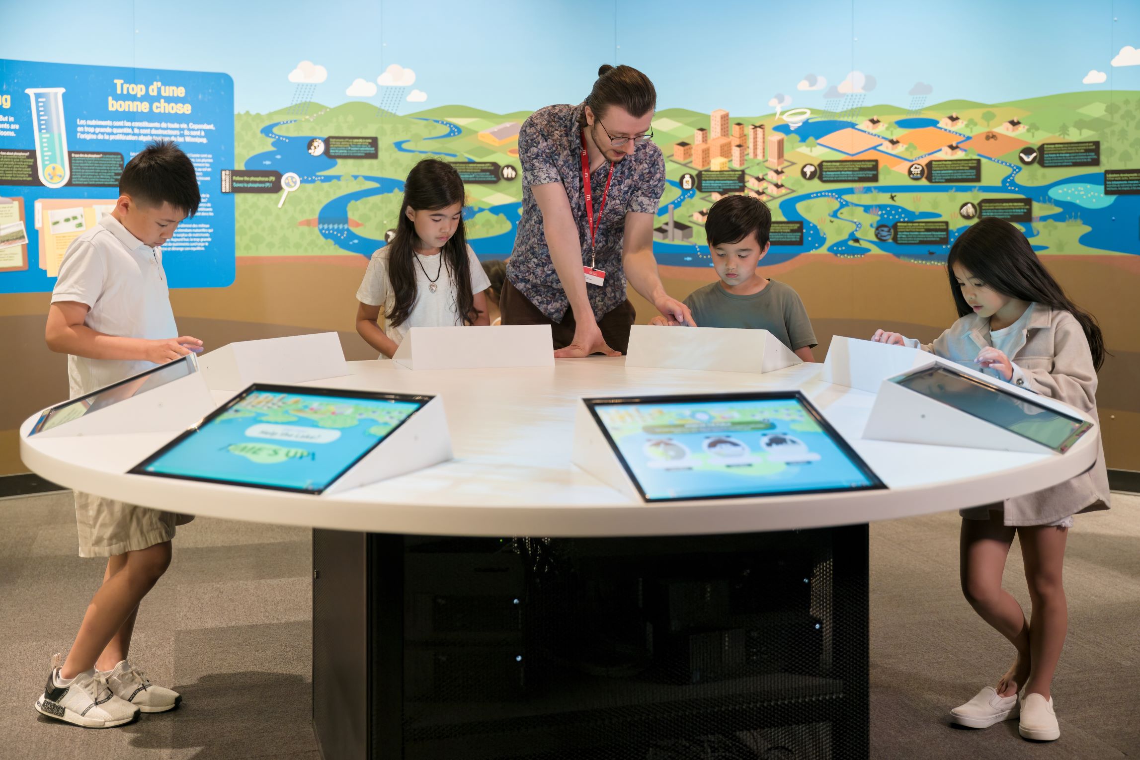 A Museum staff person standing at a round table with four children. Each child is standing at a different screen in a digital exhibit embedded in the table.