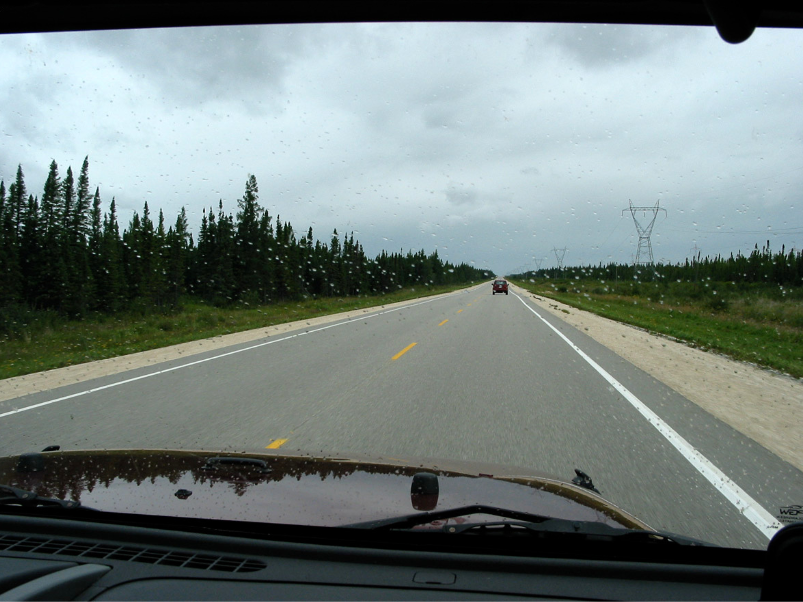 Photograph from inside a vehicle looking down a longer stretch of two-way road, with conifer trees across the ditch on either side.