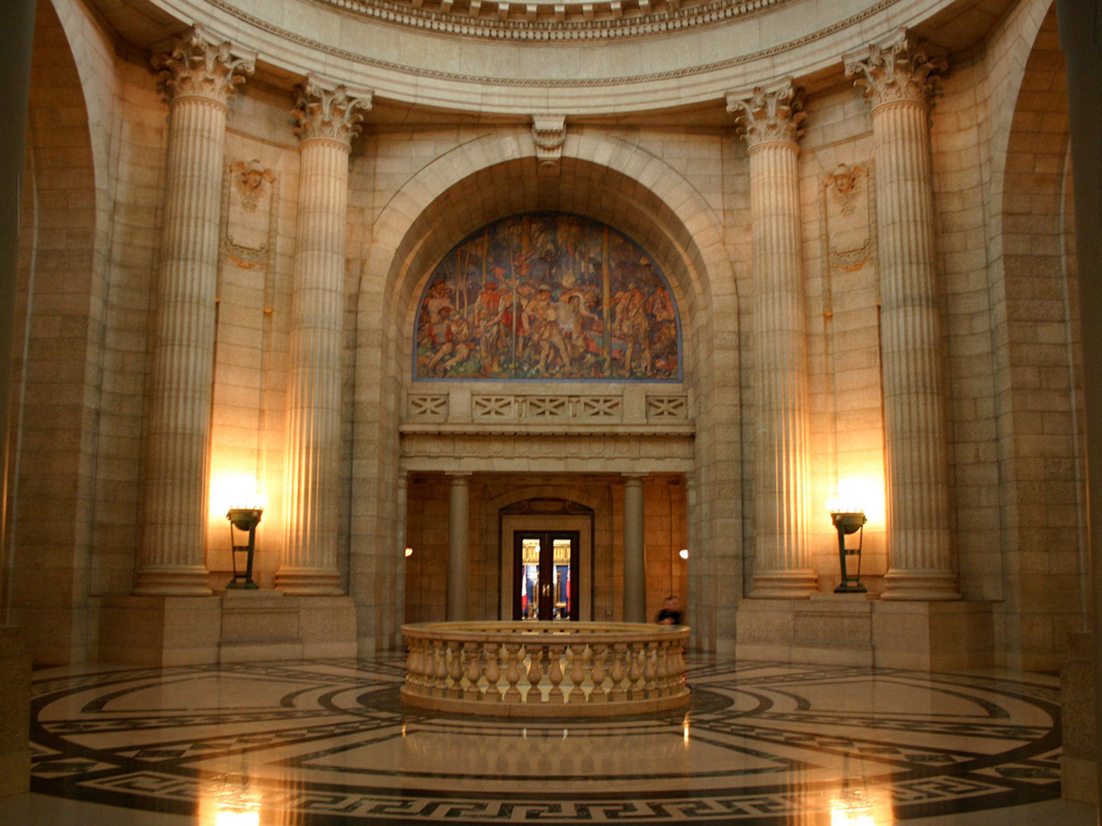 Looking towards the Rotunda in the Manitoba Legislative Building. A large foyer space with a fenced empty circling in the middle, looking down tot he floor below. Tall Tydall Stone walls with pillars encircle the space, with three arched doorways in frame.