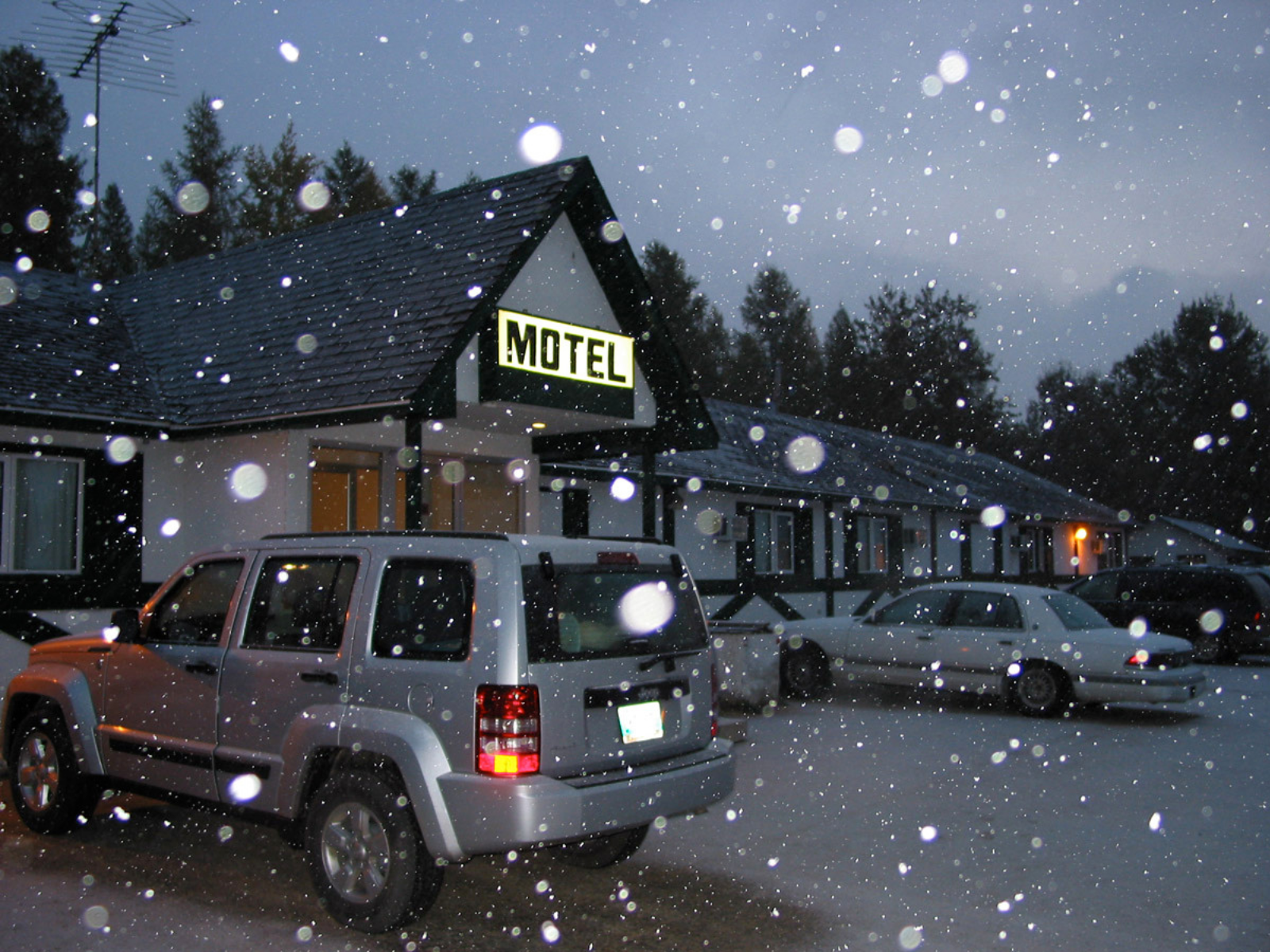 The exterior of a single-storey motels with several vehicles parked in front. Early morning light, with snow falling.