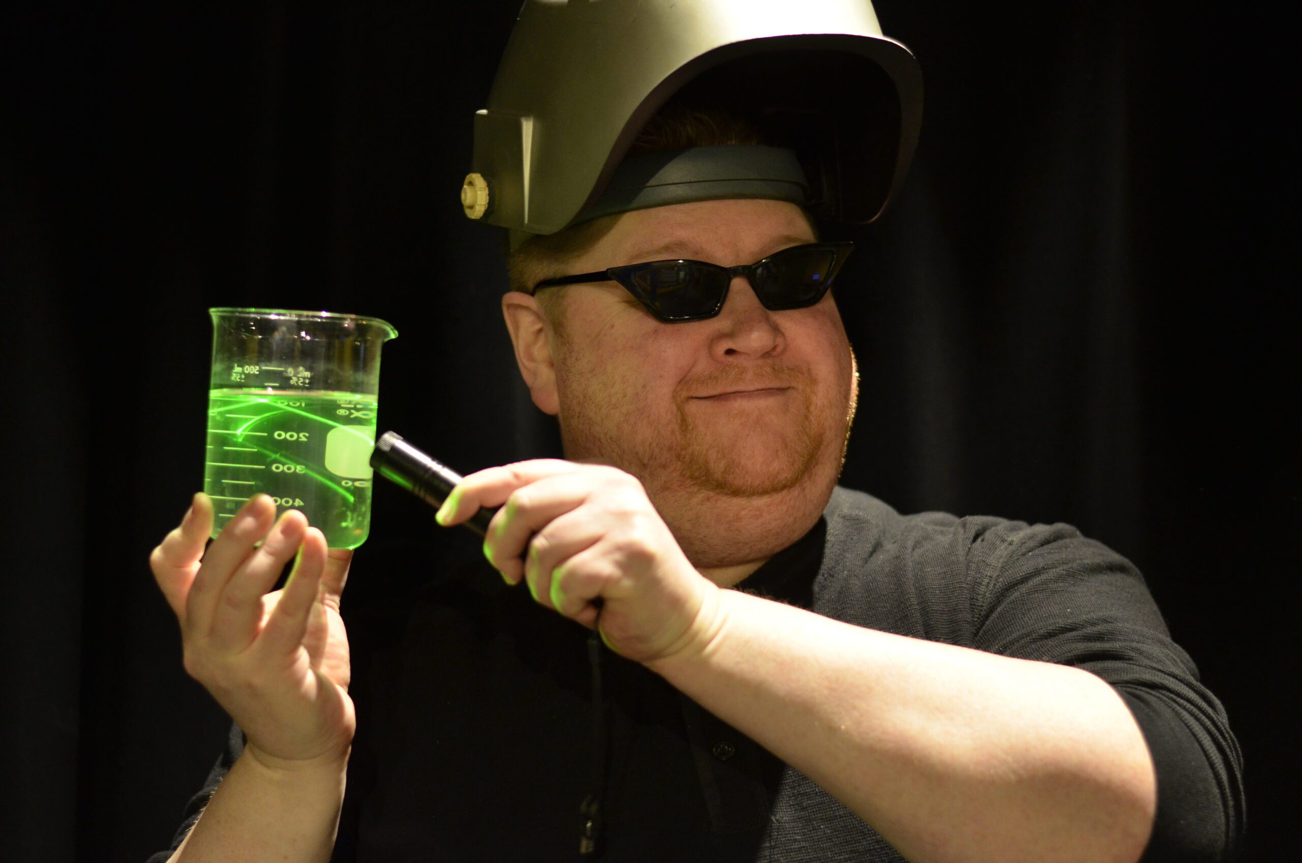 An instructor wearing a welding helmet and sunglasses holds up a laser pointer to a beaker, turning the liquid in it green as the beams reflect throughout.