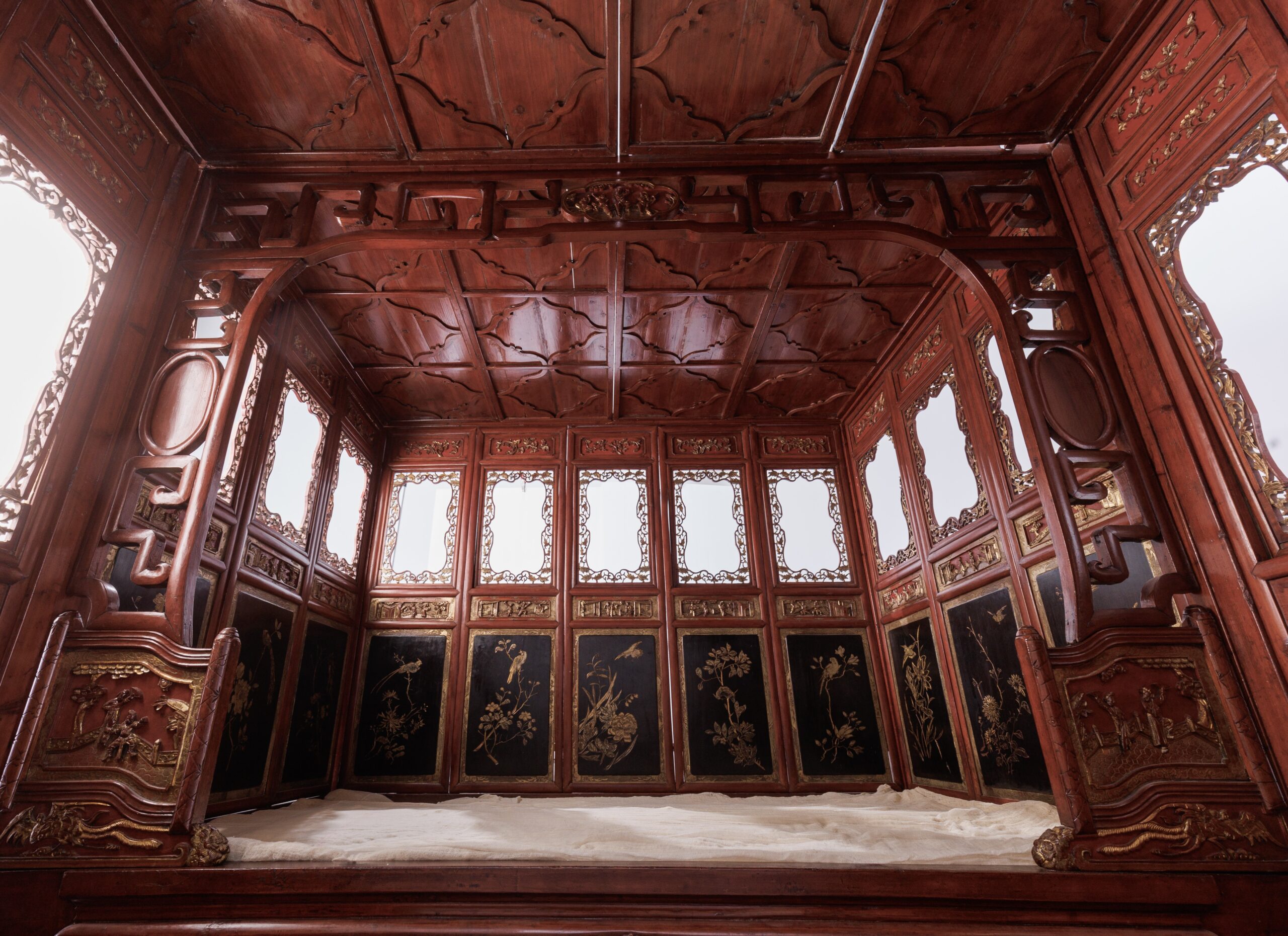 The interior of a Chinese alcove bed. The bed is entirely enclosed with a wooden ceiling, and features black and gold painted relief carvings of birds and flowers on panels around the interior.