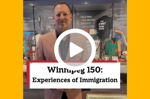 A screenshot of a video, an individual standing next to a display case designed like a kitchen table. There's a play button over the screenshot and overlaid text reads, "Winnipeg150: Experiences of Immigration".