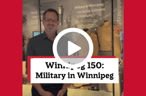 A screenshot of a video, an individual standing in front of a display case containing a number of artifacts including two army uniform jackets on dress forms. There's a play button over the screenshot and overlaid text reads, "Winnipeg150: Military in Winnipeg".