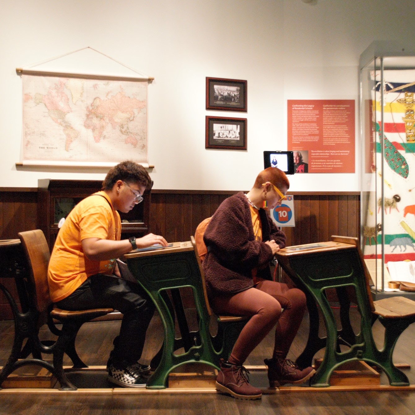 Two individuals sit at desks in the Museum's Prairies Gallery Schoolhouse exhibit, engaging with digital material embedded in the top of the desk.