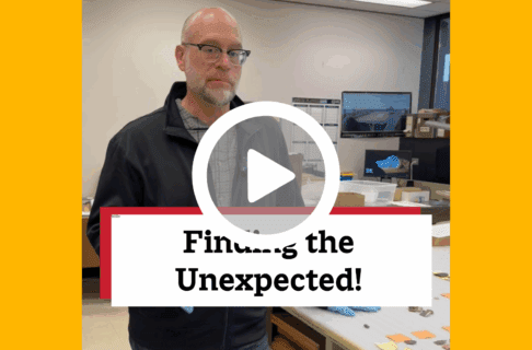 A screenshot of a video, an individual stands in a museum lab next to a table covered in artifacts. There's a play button over the screenshot and overlaid text reads, "Finding the Unexpected!".