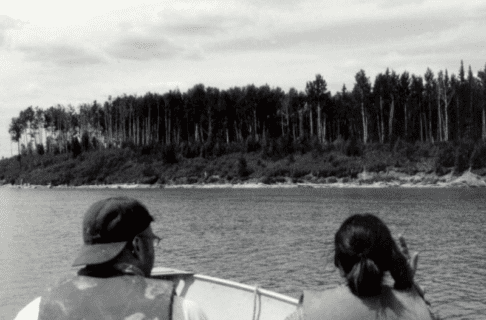 Looking over the heads of two people on a boat travelling over a body of water towards a treed bank.