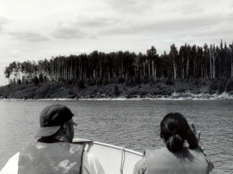 Looking over the heads of two people on a boat travelling over a body of water towards a treed bank.