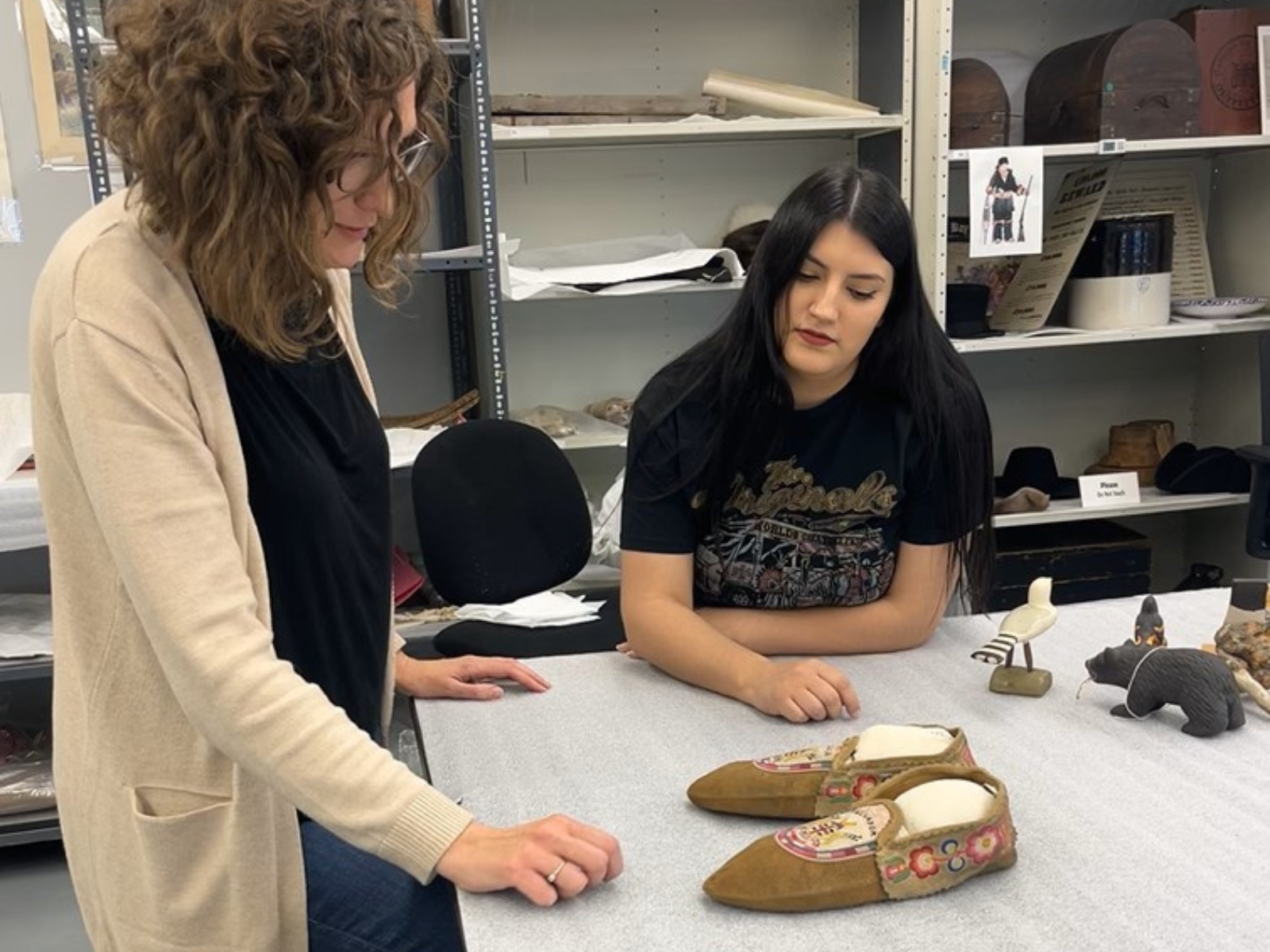 Two Museum staff at a table in a collections lab with a number of artifacts, including a pair of moccasins, laid out in front of them.