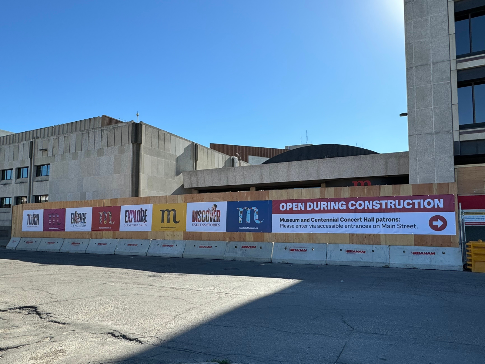 The Manitoba Museum Rupert Avenue entrance with construction hoarding in place. Branded banners with colourful Ms and a notice reading "Open during construction" and direction visitors to the Main Street entrance are affixed to the front.