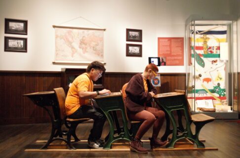 Two individuals sit at desks in the Museum's Prairies Gallery Schoolhouse exhibit, engaging with digital material embedded in the top of the desk.