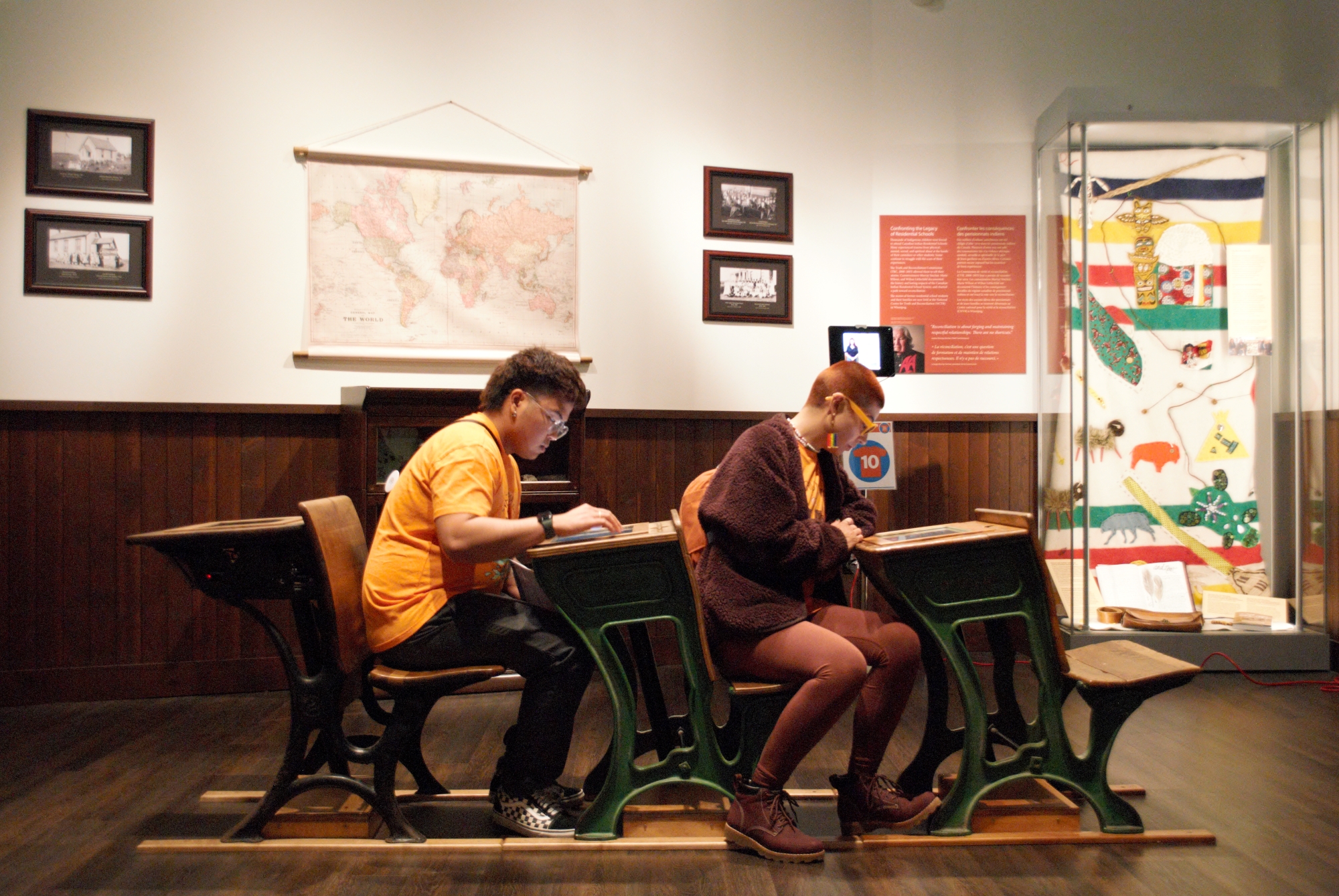 Two individuals sit at desks in the Museum's Prairies Gallery Schoolhouse exhibit, engaging with digital material embedded in the top of the desk.