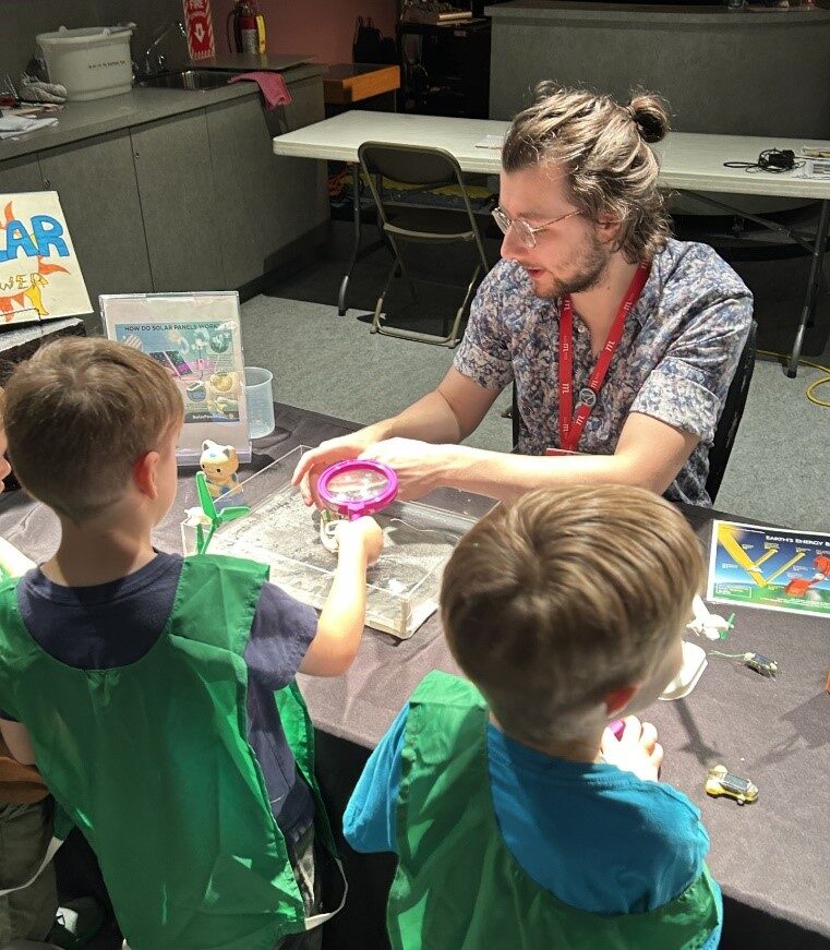 A Museum staff member seated at a programming table, demonstrating a science activity for two young visitors.