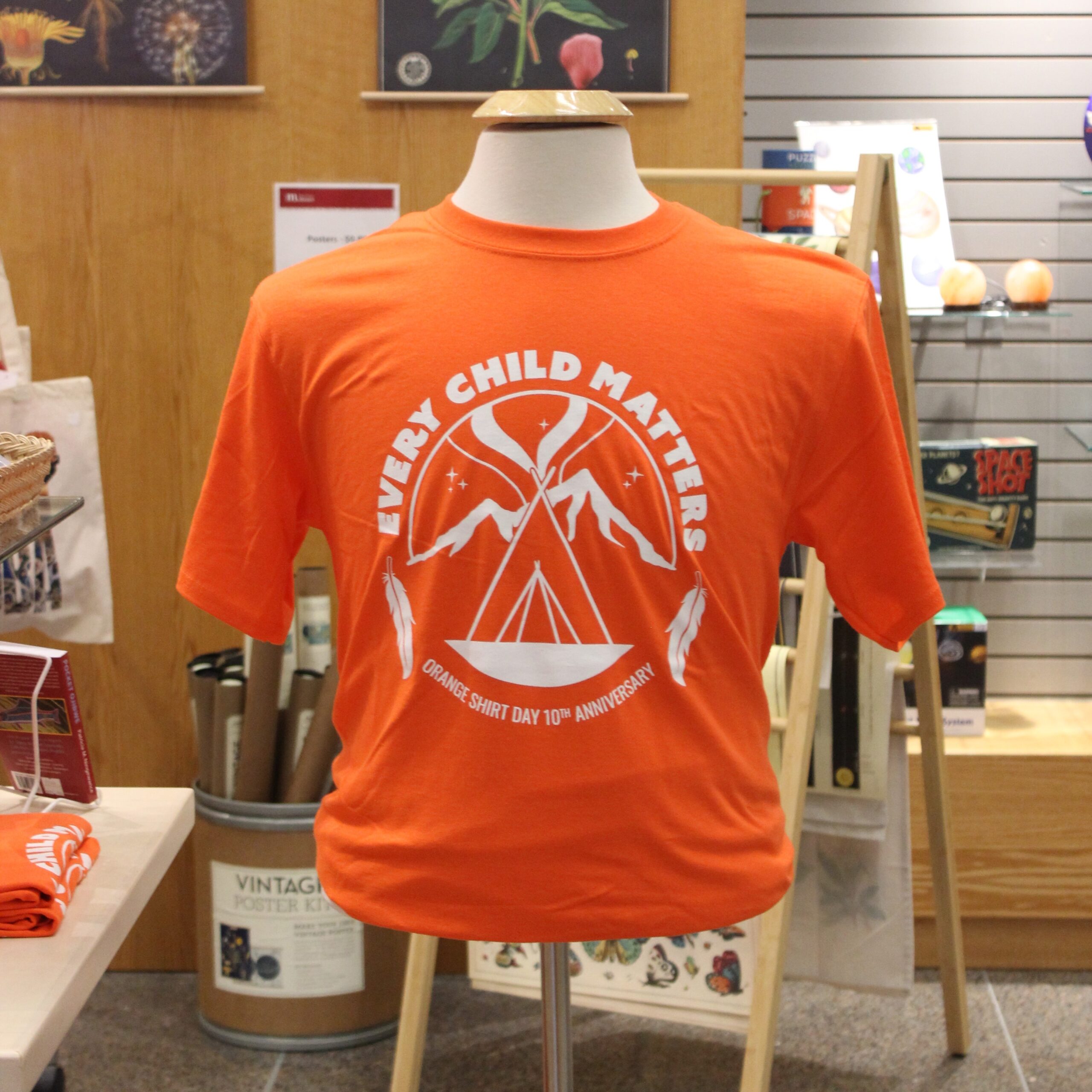 An orange t-shirt on a clothes mannequin. Design on the front features a tipi in front of mountains under the northern lights. Text around the design reads, "Every Child Matters / Orange Shirt Day 10th Anniversary".