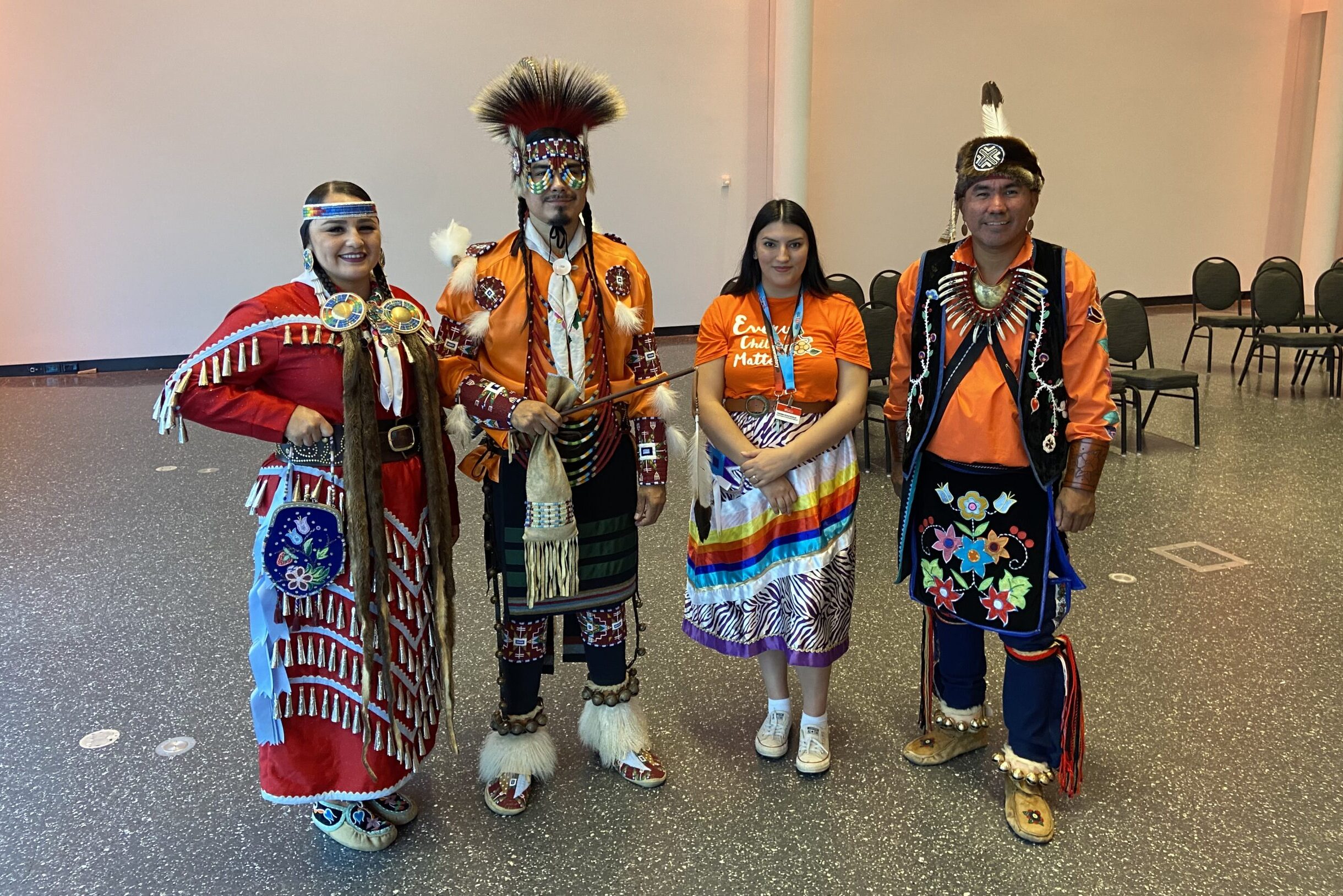 Three individuals wearing Powwow regalia stand smiling with a Museum staff member wearing an orange t-shirt and a ribbon skirt.