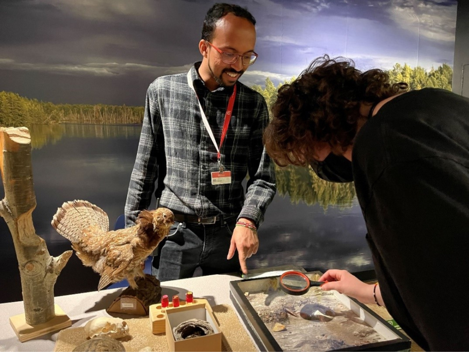 A Museum staff member behind a programming table points out features of an artifact to a visitor looking closely with a magnifying glass.