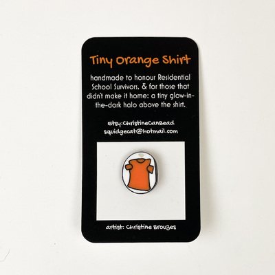 A small lapel pin featuring an orange t-shirt created by local artist Christine Brouzes.