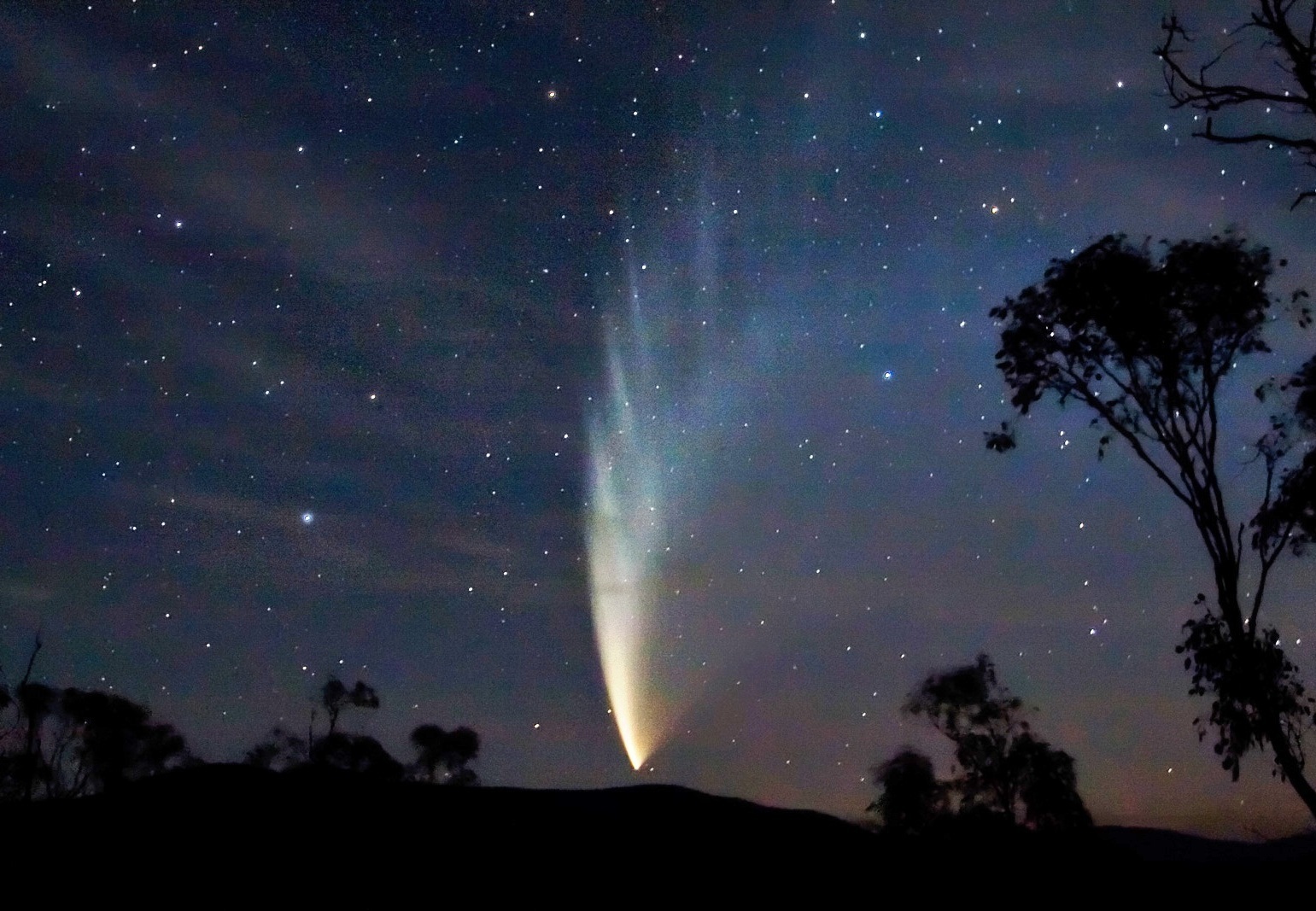 A comet with a long, bright tail streaking through the night sky, perpendicular to the ground.