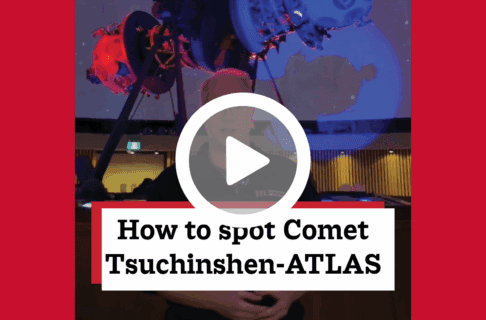 A screenshot of a video, an individual stands in front of a large Zeiss star projector in a blue and red lit room. There's a play button over the screenshot and overlaid text reads, "How to spot Comet Tsuchinshen-ATLAS".