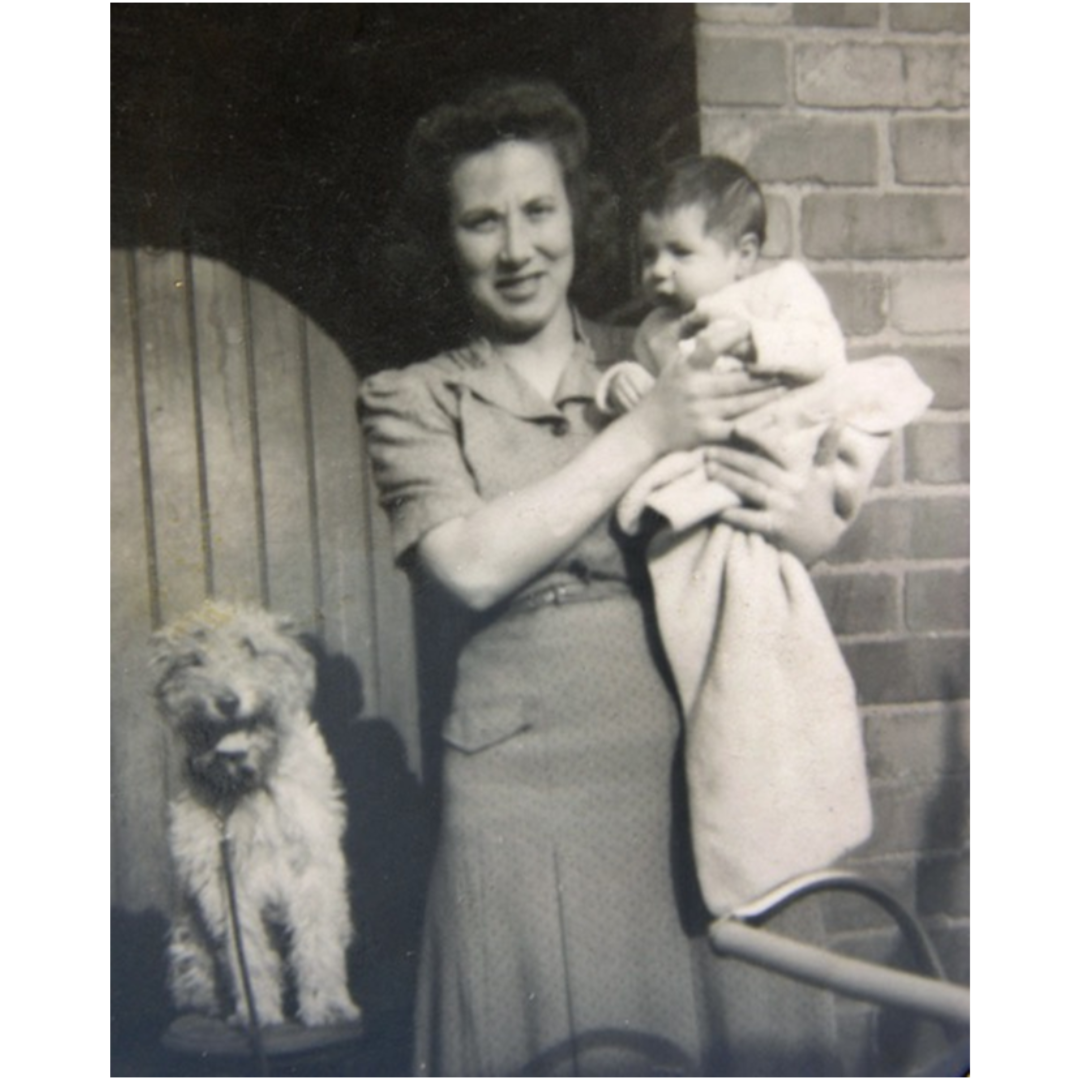 A black and white photograph of a woman standing outdoors holding a baby, with a dog seated beside them.