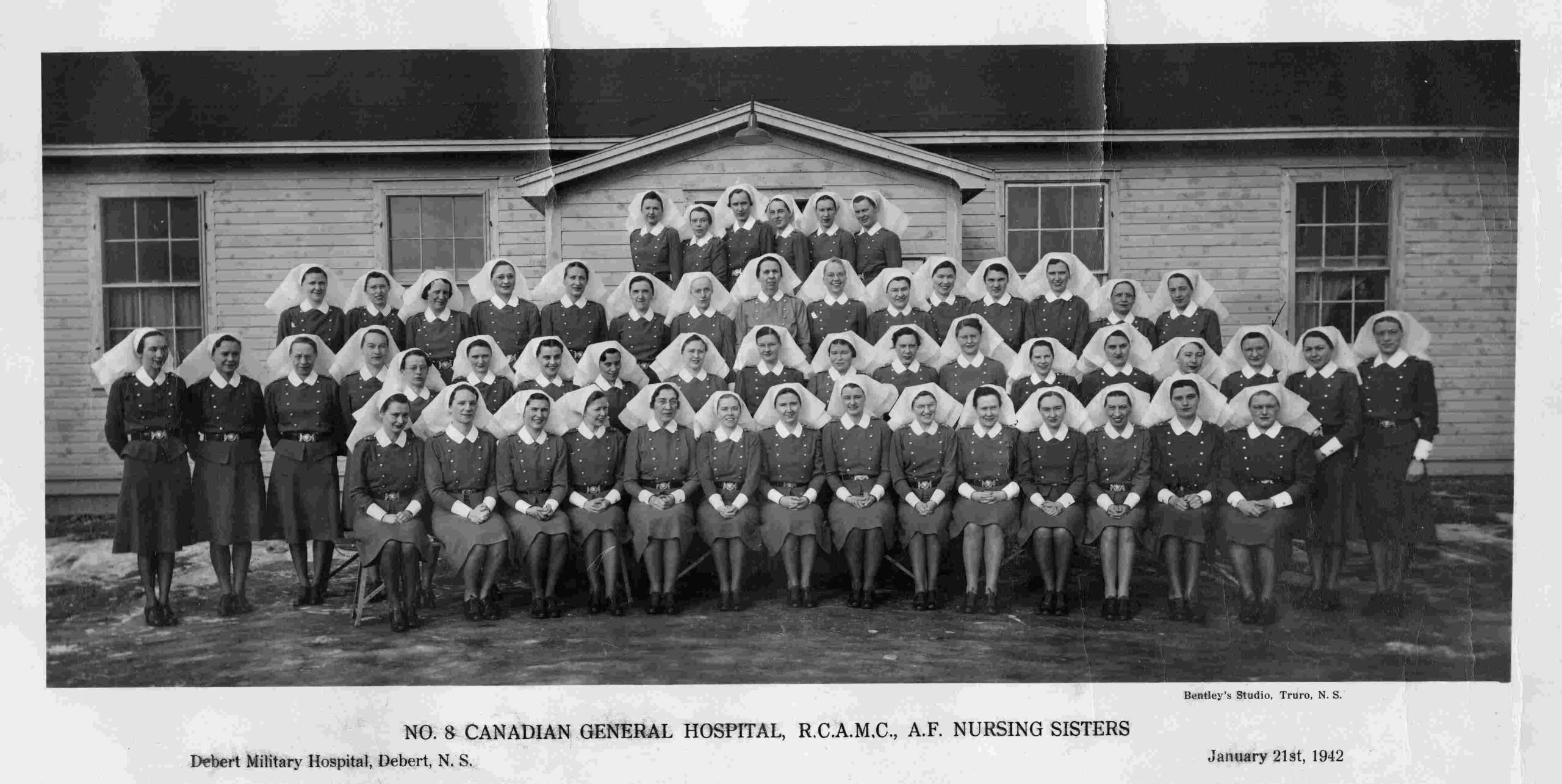 A formal group photograph of four rows of women in uniforms and nurses head scarves in front of a wooden, one-storey building.