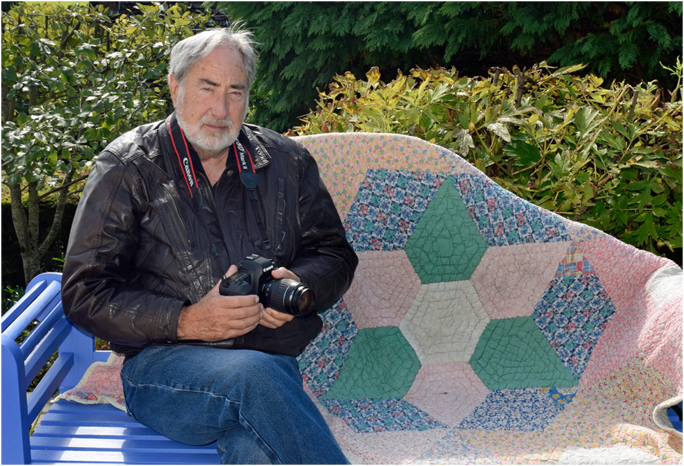 A grey-haired man holding a DSLR camera sitting on a blue bench with the quilt draped over the back of it.