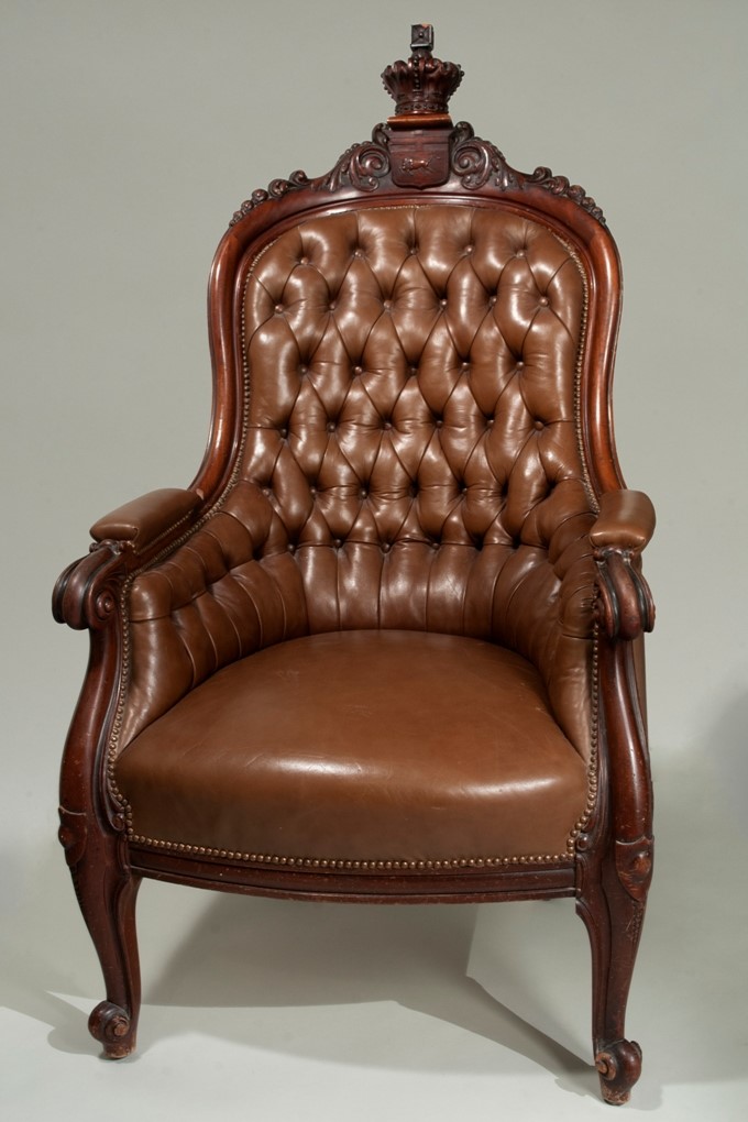 An ornate, brown leather claw-foot armchair with a wooden crest and crown embellishing the top of the seat back.