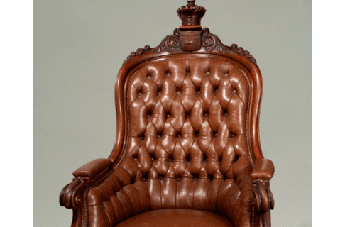 An ornate, brown leather claw-foot armchair with a wooden crest and crown embellishing the top of the seat back.