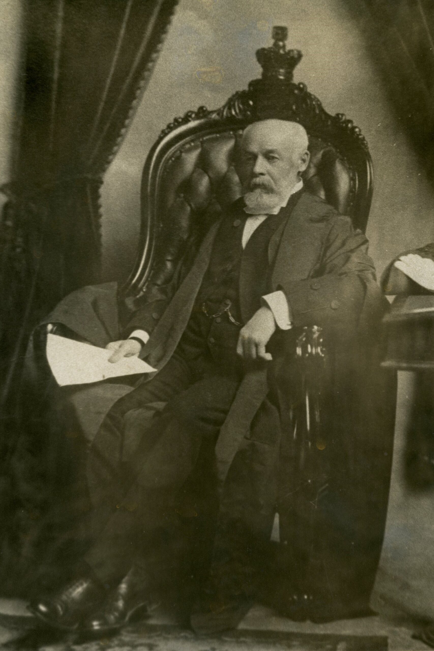 A sepia-toned photograph of a white beared man wearing a three-piece suit seated in an ornate armchair and holding papers in his right hand.