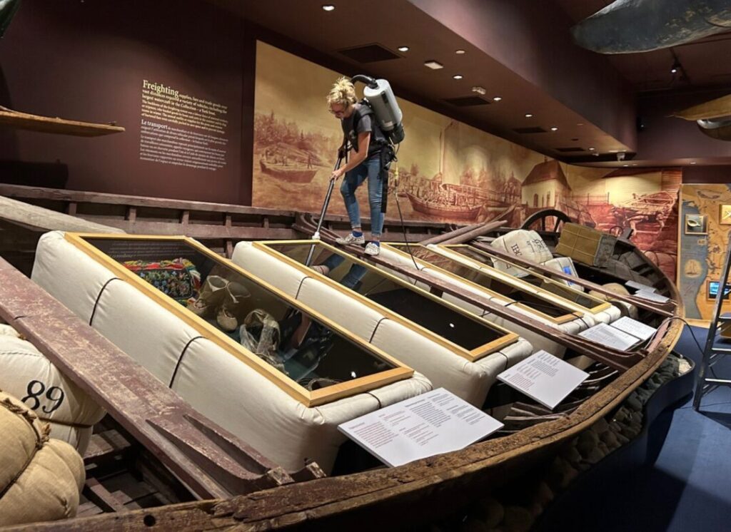 Blonde haired woman wearing blue jeans and a grey t-shirt. Woman is wearing a grey backpack vacuum and is standing on a large York boat while vacuuming an exhibit display that is inside the boat.