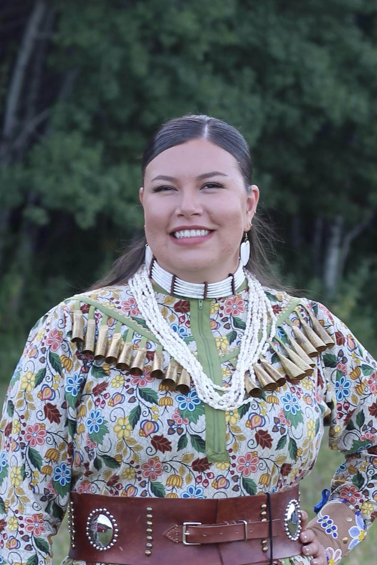 Headshot of a smiling women standing outdoors, wearing a colourful jingle dress and dentalium jewelry.