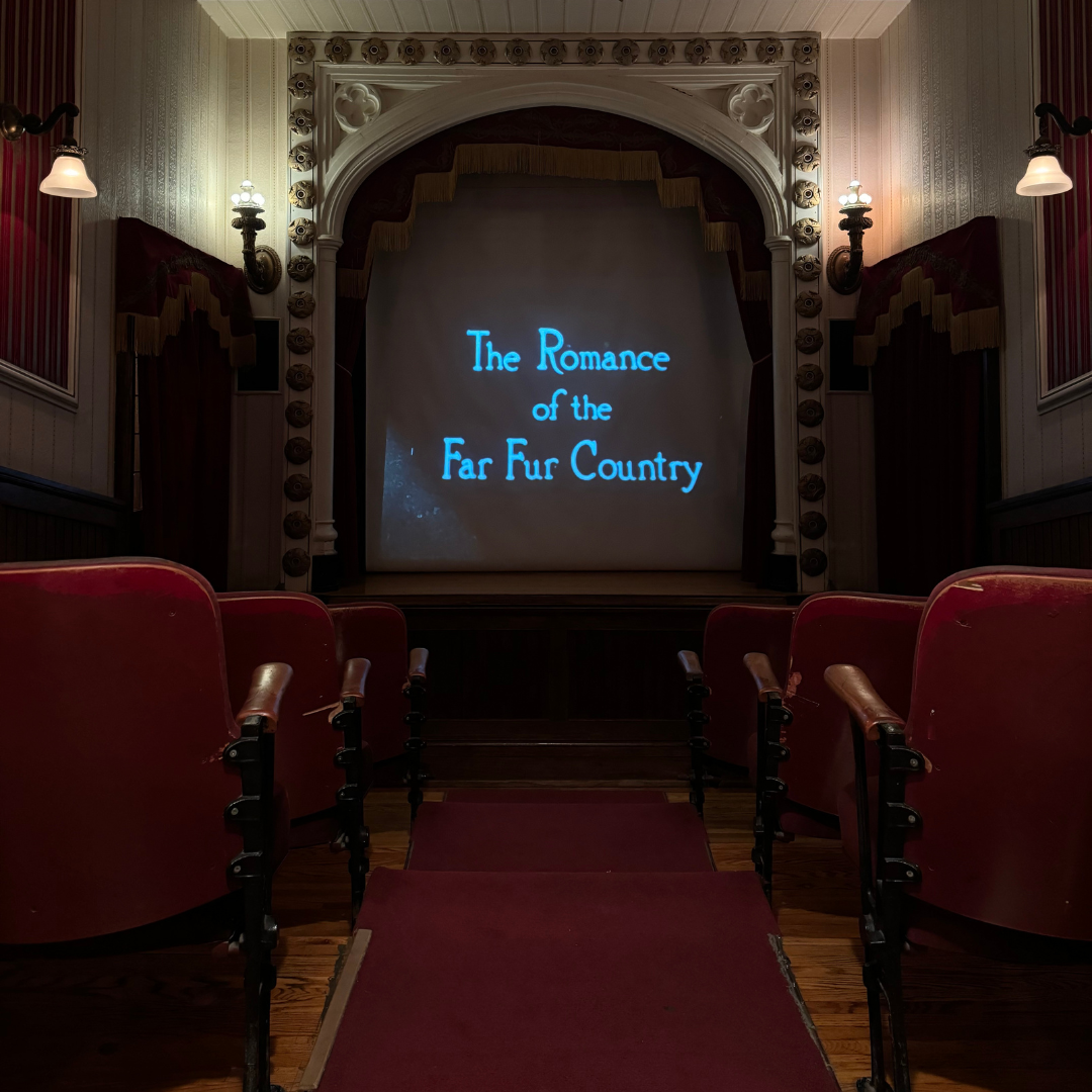 Looking down the aisle of a small theatre with red plush seats on either side. The screen is framed with detailed architecture and sconce lights. Text on the screen reads, The Romance of the Far Fur Country".