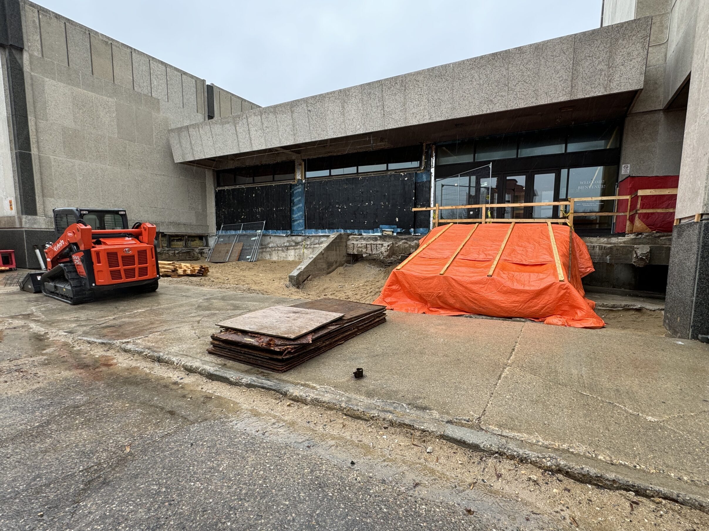 The Manitoba Museum's Rupert Avenue entrance under construction. A orange-red bobcat is parked along the sidewalk in front of a gravel area where the ramp and stairwell used to be. An orange tarp covers the previous Concourse entranceway.