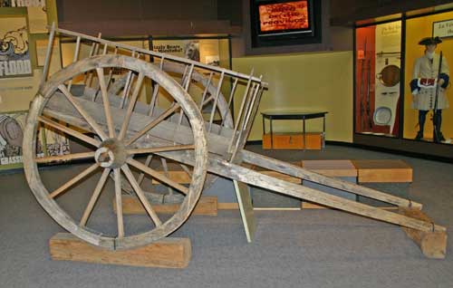 A Red River Cart on display in a museum gallery. A wooden cart with two large wheels on either side and two hitching posts stretching out in front to attach to an animal.