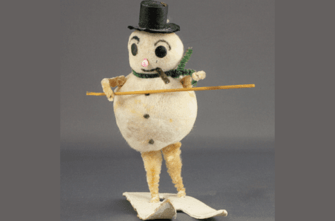 A vintage Frosty the Snowman decoration showing a snowman with legs and arms riding a set of skis while wearing a black top hat and green pipe cleaner scarf.